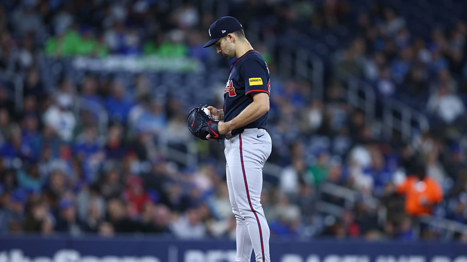Spencer Strider, an Atlanta Braves pitcher, stands on the mound holding a baseball and looking down, surrounded by fans in a blurred stadium background.